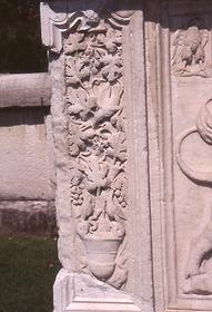 Eingesäumt wird der untere Reliefblock links und rechts von einem Weinstock, der aus einem Katharos, einem antiken Weingefäß wächst und Trauben trägt, die von Vögeln gefressen werden.