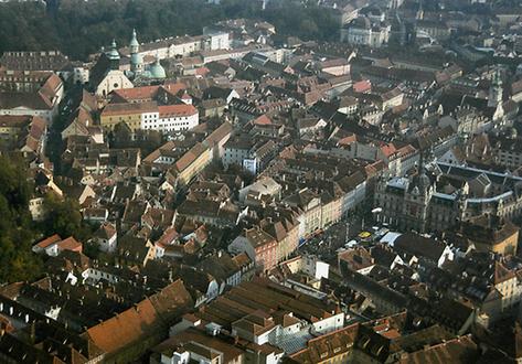 Dachlandschaft eines Teiles der Grazer Altstadt. Rechts das Grazer Rathaus, dahinter das Landhaus, links oben der Dom mit Mausoleum, davor das Priesterseminar. Ganz vorne die Dächer des Grosskaufhauses Kastner&Öhler. Die schmalen langen Satteldächer mussten auf dem vorher sichtbaren Flachdach aufgebracht werden, das vom Architektenpaar Hermann Helmer und Ferdinand Fellner Anfang des 20. Jh. konzipiert worden war. Das Flachdach störte damals dschon die örtliche Dachlandschaft und die Grazer so sehr, dass diese Änderung gefordert wurde.