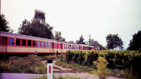 Nebenbahn der Westbahn. Eisenbahnkreuzung zwischen Lambach und Wels (Sommer 1989). Vier Schlierenwagen in rot-weißer Farbgebung und eine rote Diesellok der Baureihe 2143
