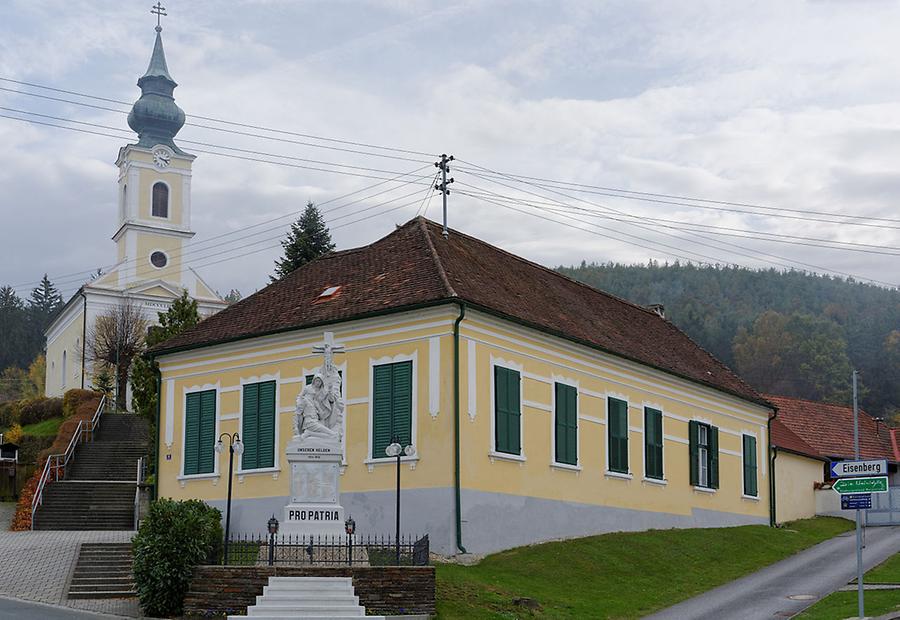 Ehemalige Schule in Badersdorf, Burgenland, Österreich, davor Kriegerdenkmal und dahinter die Filialkirche Zur Kreuzerhöhung.
