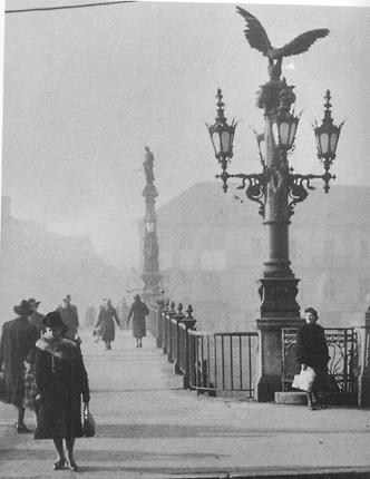 Diese Foto zeigt die nördliche Seite der 1965 abgebrochenen Hauptbrücke über die Mur in Graz. Das Foto dürfte in der Zwischenkriegszeit aufgenommen worden sein. Im Vordergrund steht eine von vier Säulen mit Adler und vier Laternen, die jeweils von Schwanenhälsen mit Flügeln gestützt werden