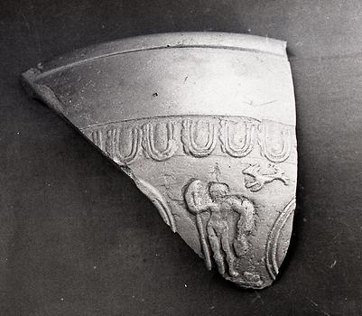 Einzelne Scherbe eines Terra-Sigillata-Gefäßes. Neben einem kurzen Streifen Banddekor erkennt man eine stehende Person und einen laufenden Hund