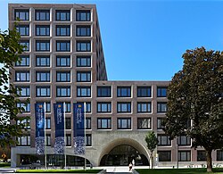 Ein Gebäude mit vielen Fenstern und rötlichbräunlicher Fassade ist zu sehen. Links bilden 10 Stockwerke einen Turm, rechts bilden fünf Obergeschoße ein Sockelgebäude. Ein großer Betonbogen führt ins Gebäude. Drei dunkelblaue Fahnen der Universität Innsbruck wehen im Vordergrund.