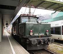 Bild der in grüner Farbe lackierten Museumslok 1020 42-6, die im Bregenzer Hauptbahnhof auf die Weiterfahrt wartet. Im Hintergrund ist der Nahverkehrstriebwagen 4024 der ÖBB zu sehen.
