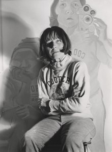  Maria Lassnig auf einem selbst fotografierten Schwarzweißbild von 1974, im Hintergrund das Gemälde Doppelselbstportrait mit Kamera