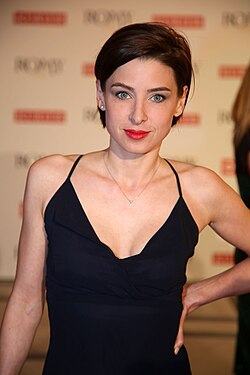 Martina Ebm bei der Romy-Verleihung 2015. Sie trägt ein ärmelloses, tief ausgeschnittenes schwarzes Kleid, das elegant und stilvoll wirkt. Ihr kurzes, dunkles Haar und rotem Lippenstift. Im Hintergrund ist ein wiederholendes Muster mit den Worten „ROMY“ und „2015“ in roter und schwarzer Schrift zu sehen. Martina Ebm trägt eine dezente Halskette mit einem kleinen Anhänger und hat eine Hand auf der Hüfte, was eine selbstbewusste Pose vermittelt.