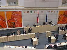 Blick von oben (wohl Zuschauerbereich) in den Parlamentsraum, große rötliche Gemälde ohne erkennbaren Inhalt, zwei Fahnen, allerlei Stühle und Tische, vereinzelt Menschen, Kurz steht als einziger mit Blick nach vorn