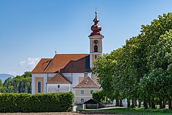 Fotografie der Kirche bei Sonnenschein und blauem Himmel. Im Vordergrund rechts ist eine auf die Kirche zuführende Kastanienallee, links im Vordergrund eine hohe Hecke. Im Hintergrund hinter der Kirche sind einige hohe Bäume zu sehen.