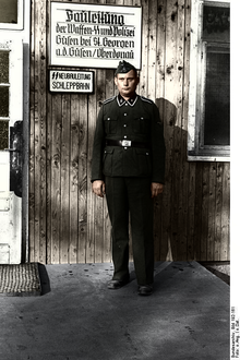 SS-Mann in Uniform vor hölzerner Baracke mit Schild "Bauleitung"