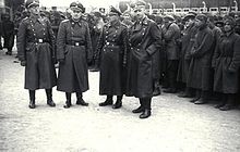 4 SS-Offiziere mit Ledermäntel und Mütze vor dutzenden schlechtgekleideten Häftlingen