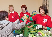 Drei freiwillige Le+O Mitarbeiterinnen in roten Leibchen im Hintergrund. Vorne eine Kundin, die von der ersten Frau Lebensmittel überreicht bekommt.