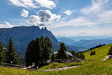 in der Bildmitte Schutzhütte von Nadelbäumen umgeben; im Vordergrund Wiese mit einem durch ein Geländer abgetrennten Weg; im Hintergrund links hohe, rechts weniger hohe Berge