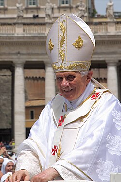 Farbfotografie vom Papst in liturgischer Kleidung und Papstkrone aus Stoff mit goldenen und roten Ornamenten. Im Hintergrund sind ein altes Gebäude und Menschen zu sehen.