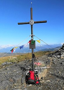 mehrere Meter großes Gipfelkreuz aus Metall; mit Seilen abgespannt; an diesen hängen teilweise verwitterte Wimpel etc.; vor dem Gipfelkreuz ein Rucksack; im Hintergrund Teile eines Steinstapels zu sehen; in der Entfernung grasbedeckte Hochebene; dahinter weitere Gipfel mit Fels und Schnee