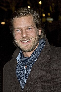 Henning Baum trug bei der Berlinale 2008 ein elegantes, klassisches Outfit. Trenchcoat Mantel, blaues Hemd, bei dem nur der Kragen sichtbar ist, darüber mit einem locker gebundenen Halstuch und kurzrasiertem Vollbart.