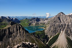 Luftaufnahme: zwischen zwei Berggipfel liegt ein Stausee mit grün-blauen-Wasser; am Rand des See ein Gebäudes; aufgrund der Entfernung kaum näher erkennbar