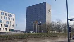 Moderne städtische Szene mit zwei markanten Gebäuden, die zusammen ein Büro- oder Verwaltungszentrum darstellen: Rechtes Gebäude: Es ist ein hohes, rechteckiges Bauwerk mit einer grauen Fassade, die in klaren Linien gestaltet ist. Die Fenster sind in mehreren horizontalen Reihen angeordnet. Oben rechts an der Fassade befindet sich ein auffälliges, goldfarbenes Logo in der Form eines stilisierten „G“. Linkes Gebäude: Dieses Bauwerk ist kleiner und hat eine weiße Fassade mit großen Fenstern, die in einem gleichmäßigen Raster angeordnet sind. Es wirkt offen und modern, im Gegensatz zum eher massiven Stil des rechten Gebäudes und hat einen gelben Balkon.Aglaia Konrad entwarf den weithin sichtbaren gelben Balkon am Provinzhaus der Provinz Flämisch-Brabant. Umgebung: Im Vordergrund sind fünf Fahnenmasten mit verschiedenen Flaggen zu sehen. Darunter befindet sich die belgische Nationalflagge sowie andere regionale oder organisatorische Flaggen. Die Umgebung ist von einem klaren Himmel mit blauem Hintergrund geprägt, und im unteren Bereich des Bildes sind einige kahle Bäume und ein gepflegter Grünstreifen sichtbar