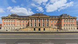 Frontale Farbfotografie eines roten Schlosses mit zwei Stockwerken und braunen Säulen. Drei stark verzierte Wappen hängen am Gesims und zwei Seitentreppen führen über eine Mauer zum Eingang. An der Mauer befinden sich drei Türen. Über dem Eingang und den vier Fenstern des Mittelteils hängen Kopfreliefs. Im Vordergrund sind eine Straßenbahnschiene und eine Straße zu sehen.