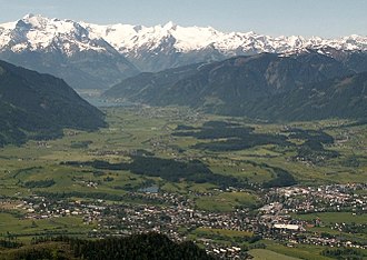 Saalfeldener Becken (Blickrichtung Süden) mit Kühbichl und Kollingwald hinter dem Ritzensee, vorn die Stadt Saalfelden, rechts der Biberg und die bewaldeten Ausläufer gegen den Zeller See, hinten Hohe Tauern mit Wiesbachhorn (links) und Kitzsteinhorn (rechts)