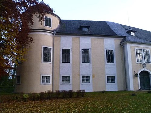 Links ein dreigeschoßiger, runder Eckturm, rechts ein Torbau mit Korbbogentor und gekuppeltem Fenster, dazwischen ein zweigeschoßiger Trakt mit drei Achsen und beige-weißem Anstrich