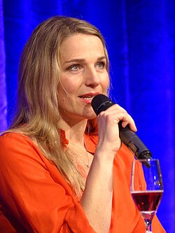 Tanja Wedhorn mit langen  Haaren spricht in ein schnurloses Stabmikrofon, welches sie mit der rechten Hand hält. Sie trägt ein orange-rotes Oberteil und eine Kette mit einem Anhänger. Sie sitzt vor einem blauen Vorhang  und vor ihr seht ein halb geleertes Glas mit Rotwein.