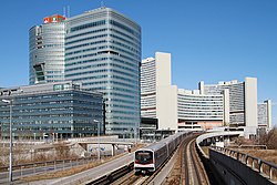 Zug der Type V auf der U1 vor der Skyline der Donau City; im Hintergrund die Station Kaisermühlen – Vienna International Centre