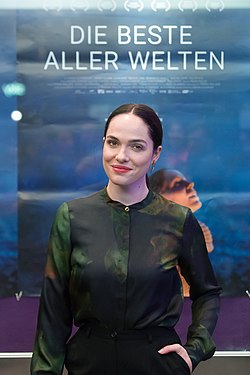 Verena Altenberger mit dunklen Haaren steht vor einem Filmplakat. Auf dem Plakat ist der Titel "DIE BESTE ALLER WELTEN" in großen weißen Buchstaben oben zu lesen. Der Hintergrund des Plakats hat einen Farbverlauf von Blau zu Lila mit einigen verschwommenen Formen und Texten. Sie trägt ein dunkles, elegantes, langärmeliges Hemd mit einem dezenten Muster und schwarze Hosen. Das Bild wurde bei der Filmpremiere für den Film "Die Beste Aller Welten" in Wien 2017 aufgenommen.